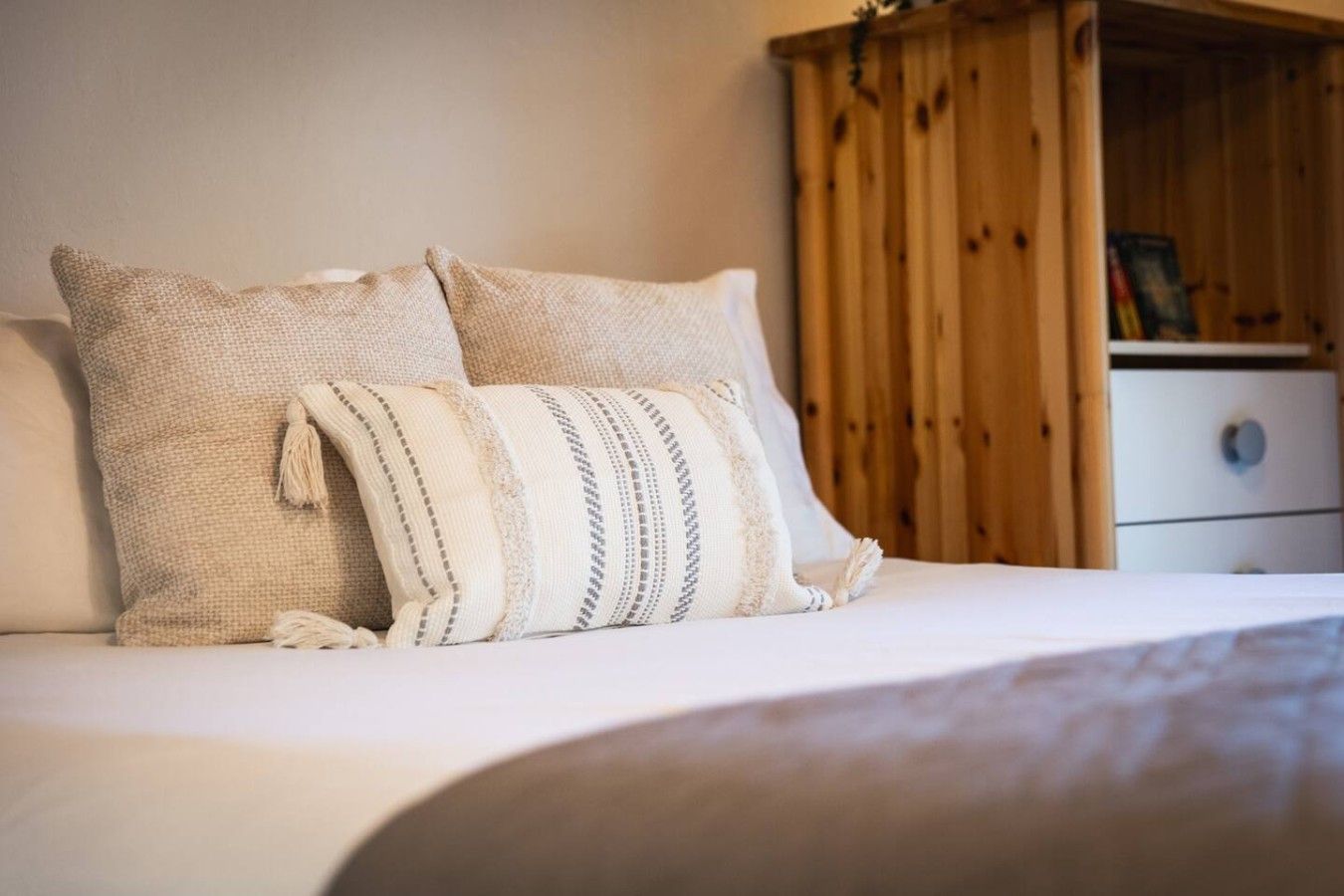 A bed with two pillows and a blanket, a wooden wardrobe, and a white bedside table.