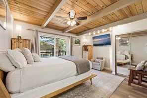 Quarto com cama grande, vigas de madeira no teto, ventilador de teto e vista através de uma porta de vidro deslizante.