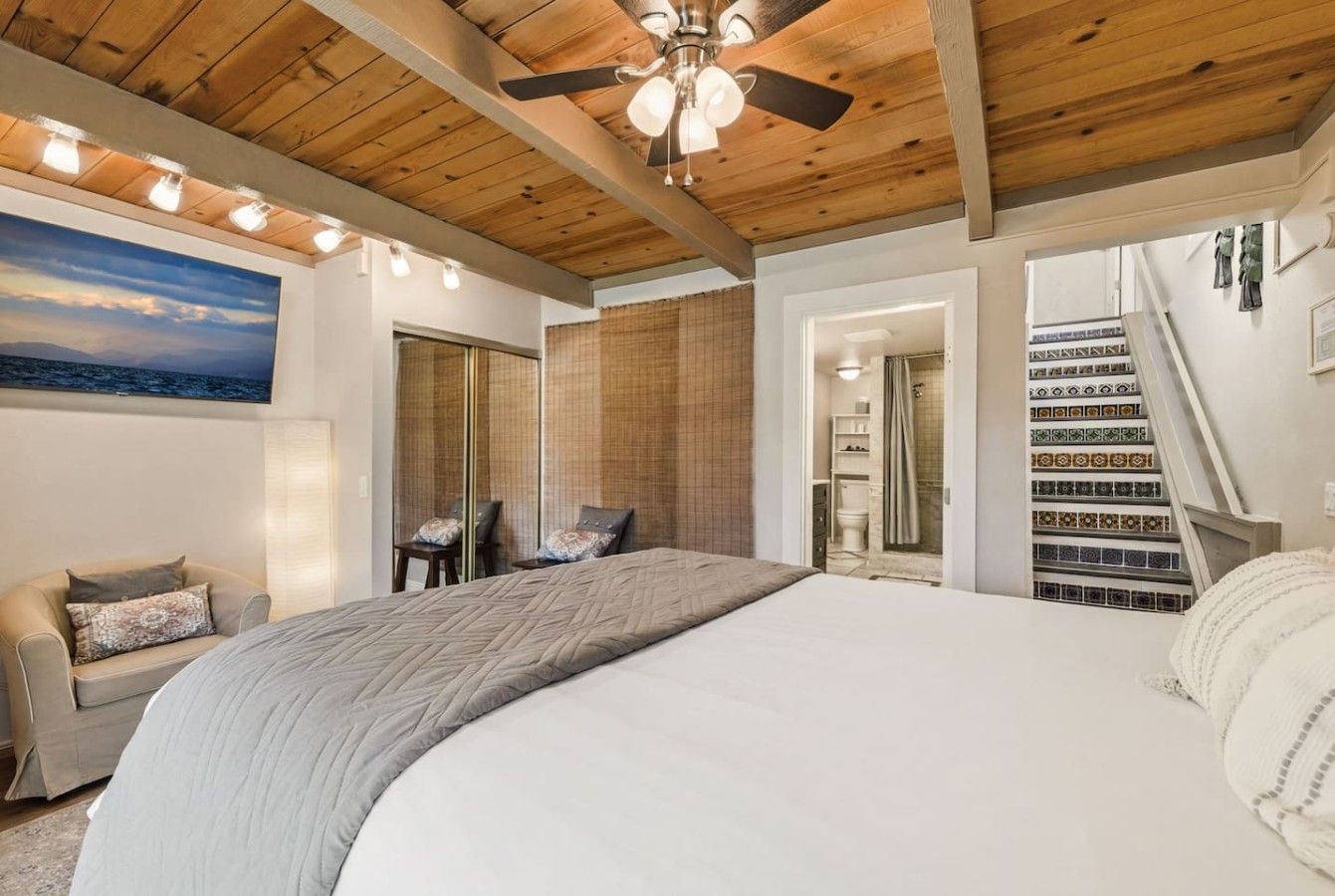A bedroom with a large bed, a ceiling fan, a view of the sea through a window, and a staircase leading to an upper level.