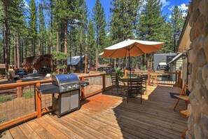 A wooden deck with a grill, outdoor table, and umbrella, surrounded by tall pine trees.