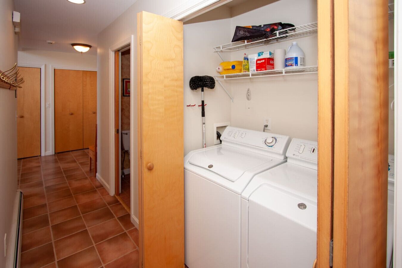 A laundry room with a washing machine and dryer, a shelf with cleaning supplies, and a door leading to another room.
