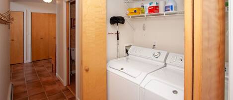 A laundry room with a washing machine and dryer, a shelf with cleaning supplies, and a door leading to another room.
