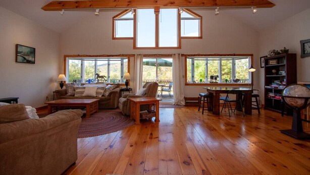 A spacious living room with wooden floors, a high ceiling with exposed beams, a dining area with a table and chairs, and large windows offering a view of the outdoors. - Killington Retreat | Deck-Fire Pit-Mountain Views! (Clarendon)