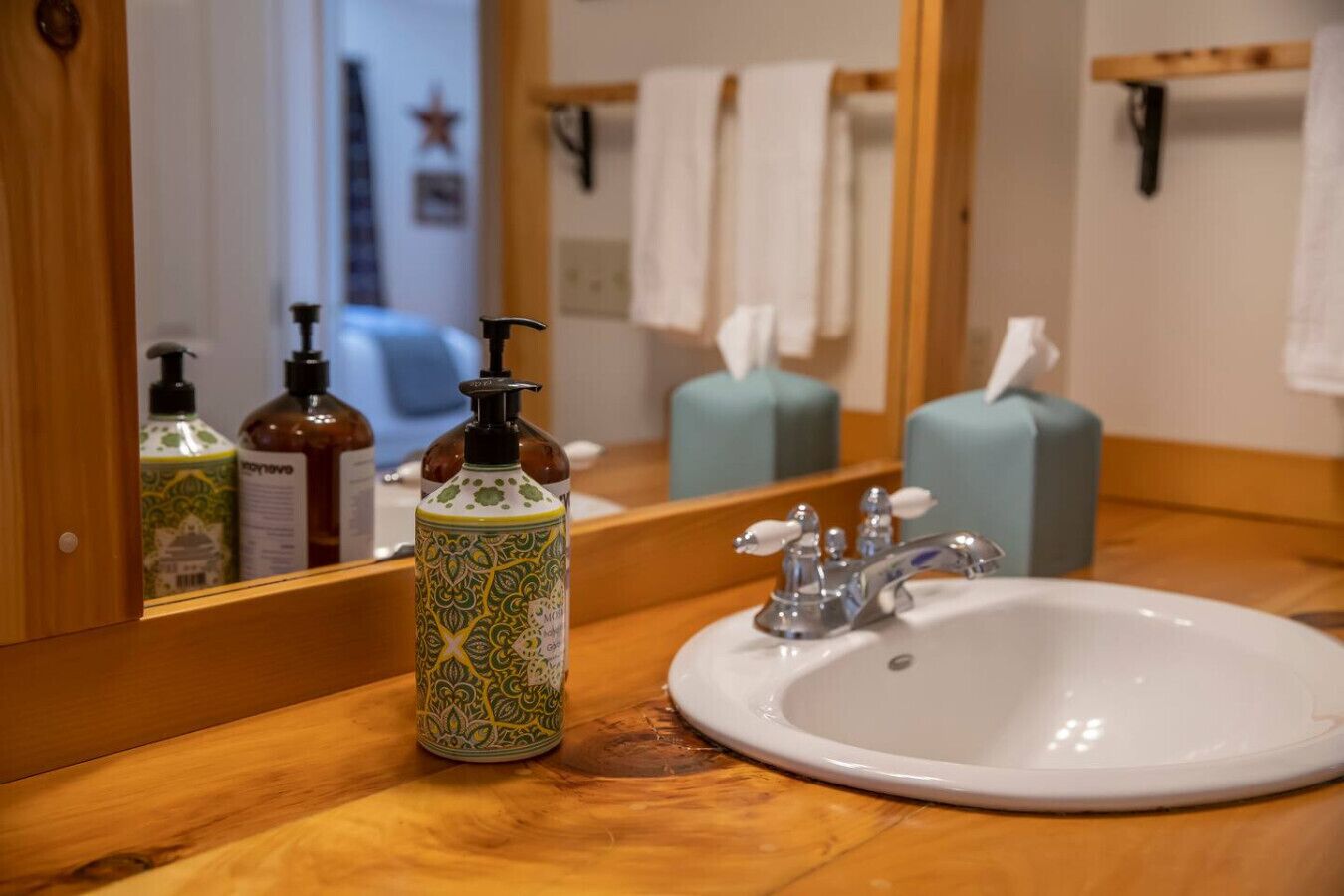 A bathroom with a wooden vanity, a white sink, a mirror, and toiletries including a patterned bottle and a pump bottle.