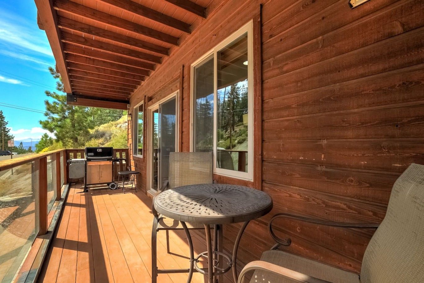A wooden deck with a round table and chairs, a grill, and large windows.