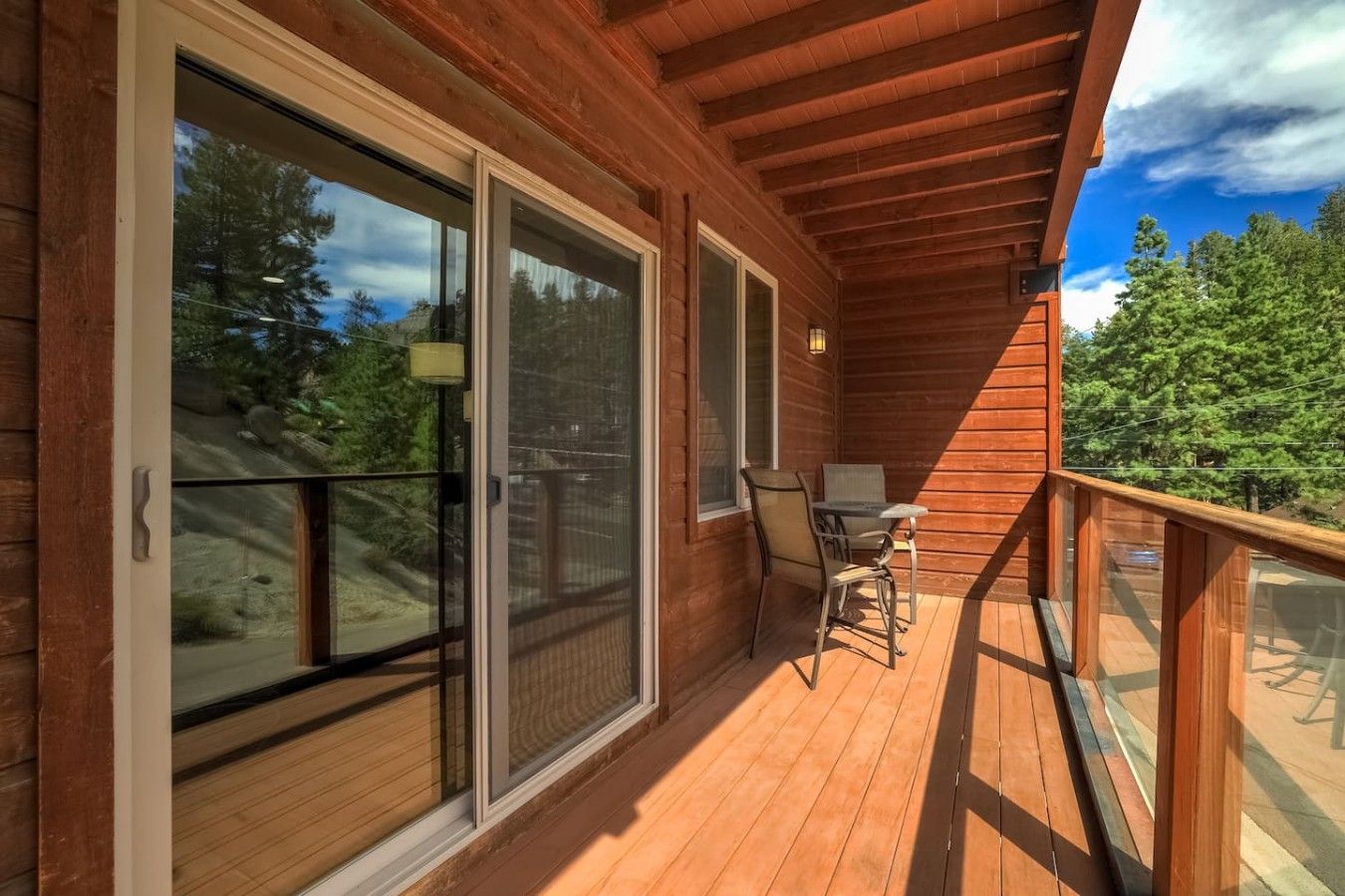 A wooden deck with a table and chairs, large sliding glass doors, and a view of trees.