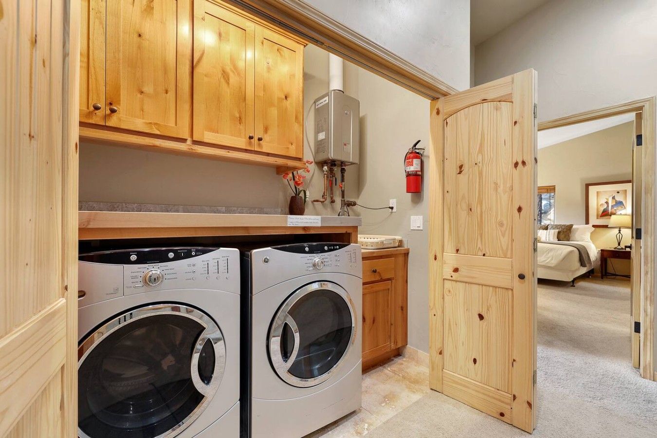 A laundry area with two front-loading washing machines, wooden cabinets, a fire extinguisher, and a door leading to a bedroom.