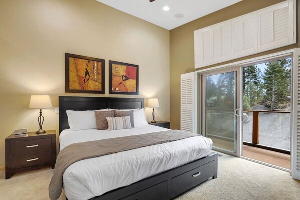 A bedroom with a bed, two bedside tables, a nightstand, a sliding glass door to a balcony, and two wall-mounted paintings.