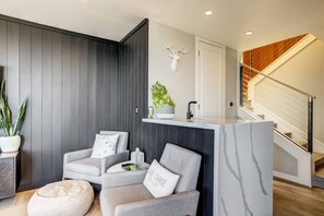 A modern living room with a grey sofa, a small round ottoman, a white wall-mounted shelf with a plant, and a wooden staircase.