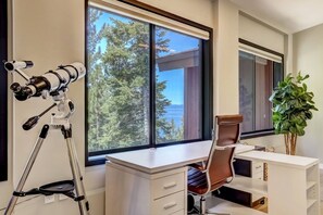A modern office with a desk, chair, telescope, and large windows overlooking a forested area.