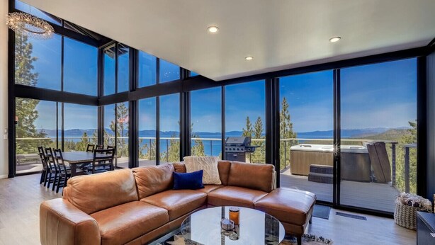 A modern living room with a leather sectional sofa, a glass coffee table, and a dining area with a table and chairs. Large windows offer a view of the outdoors. - Luxury Estate | Hot Tub & Lake Views | Sleeps 8 (Glenbrook)