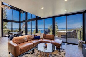 A modern living room with a leather sectional sofa, a glass coffee table, and a view of a lake and mountains through large windows.