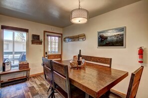 Een eetgedeelte met een houten tafel, stoelen, een hanglamp, een ingelijst schilderij en een brandblusser.