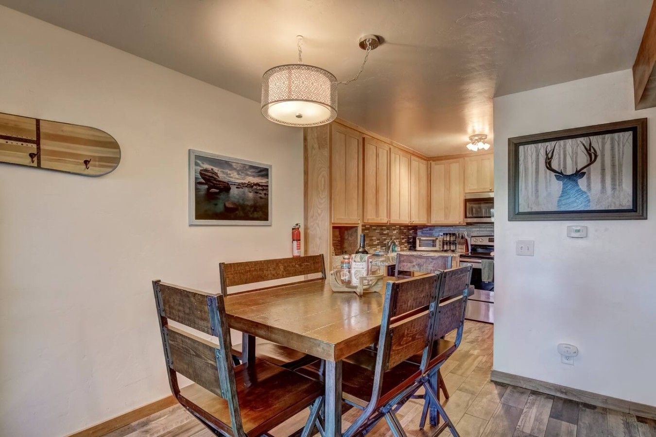 A dining area with a wooden table and chairs, a hanging light fixture, and kitchen cabinets in the background.