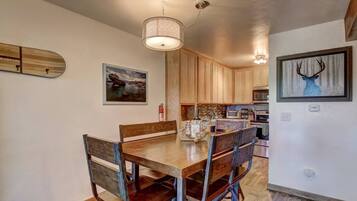 A dining area with a wooden table and chairs, a hanging light fixture, and kitchen cabinets in the background.