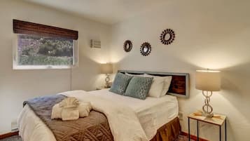 Cama bem arrumada com colcha, travesseiros e roupão. Duas luminárias de cabeceira e uma janela com vista para árvores.