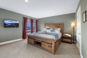 Une chambre à coucher avec un lit en bois, une table de chevet, une lampe, une fenêtre avec des rideaux et un téléviseur fixé au mur.