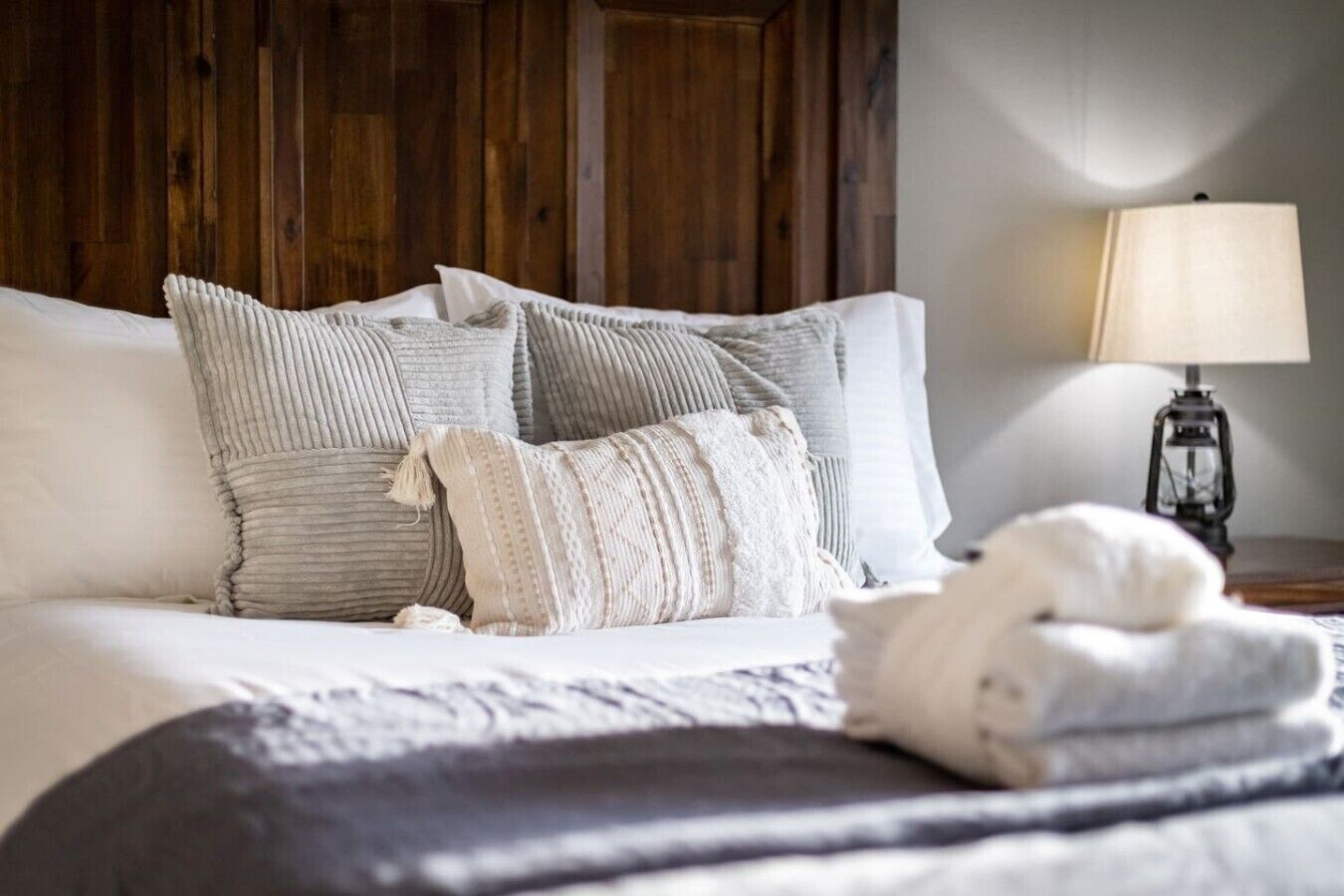 Uma cama com várias almofadas e um cobertor, uma luminária em uma mesa de cabeceira e uma cabeceira de madeira.