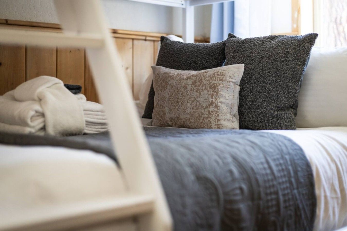 A bed with a grey and white patterned bedspread, a wooden headboard, and a white chair with a folded blanket.