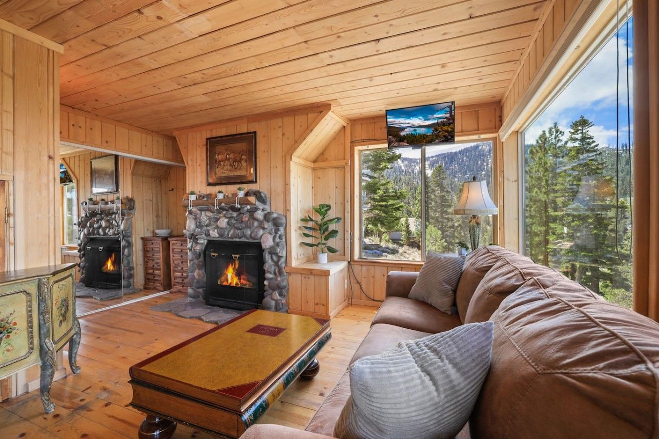 Ein gemütliches Blockhaus-Interieur mit Kamin aus Naturstein, Holzboden und einem großen Fenster mit Blick auf den Wald.