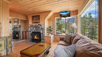 Interior acogedor de una cabaña con chimenea de piedra, techo de madera y un ventanal con vistas al bosque.