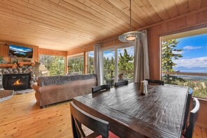 A cozy cabin interior with a stone fireplace, a sofa, a dining table, and large windows offering a scenic view. - Heavenly Escape | Lake Views, Pets OK | Sleeps 6 (Stateline)