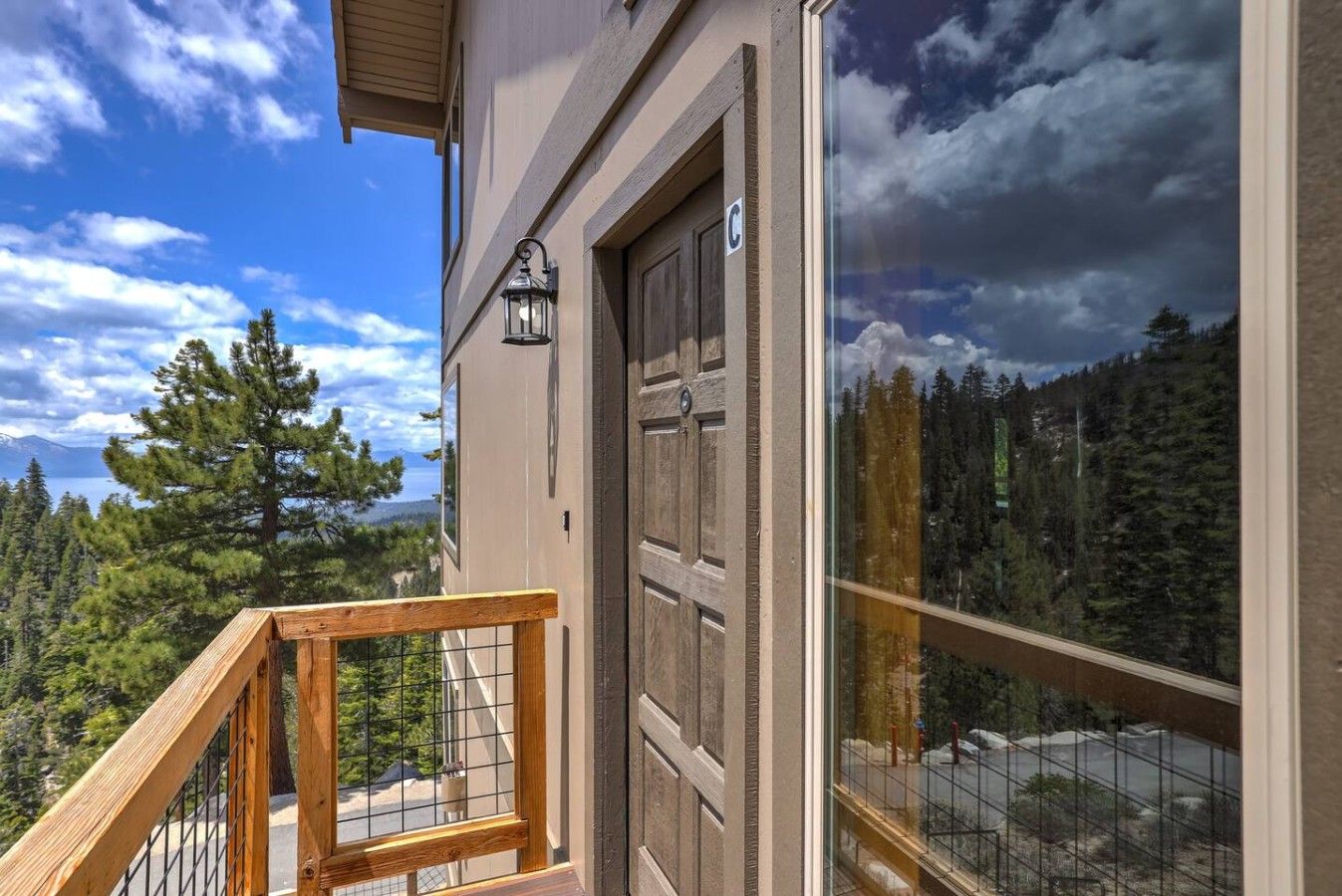 A wooden balcony with a metal railing, a door with a window, and a view of a forested landscape.