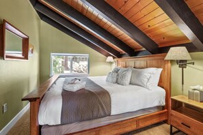 Un dormitorio con techo inclinado, una cama con ropa de cama blanca, una mesita de noche de madera y un ventanal con vistas a árboles.