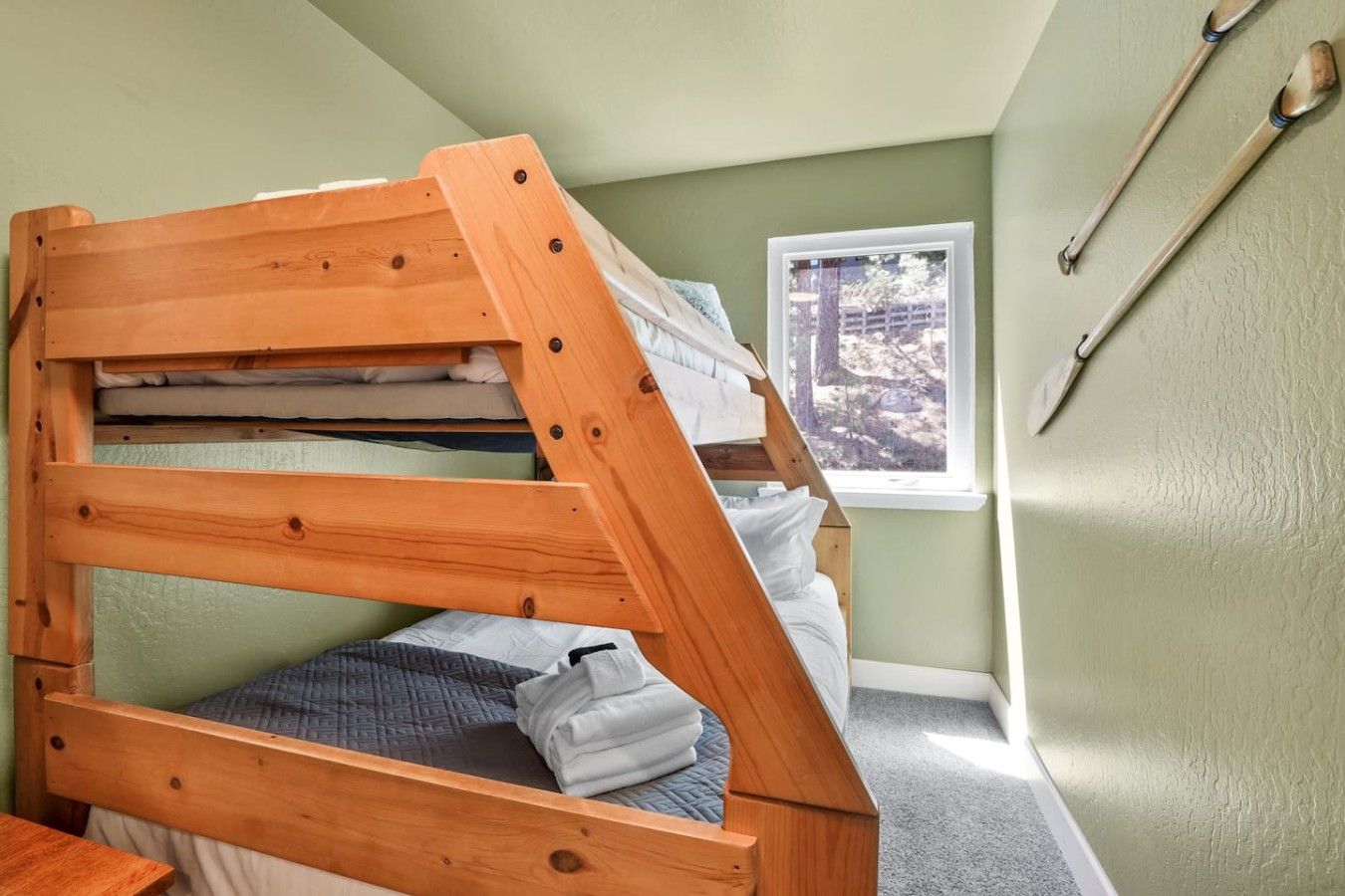 A bunk bed with a wooden frame, a window with a view of trees, and a wall-mounted wooden spoon.