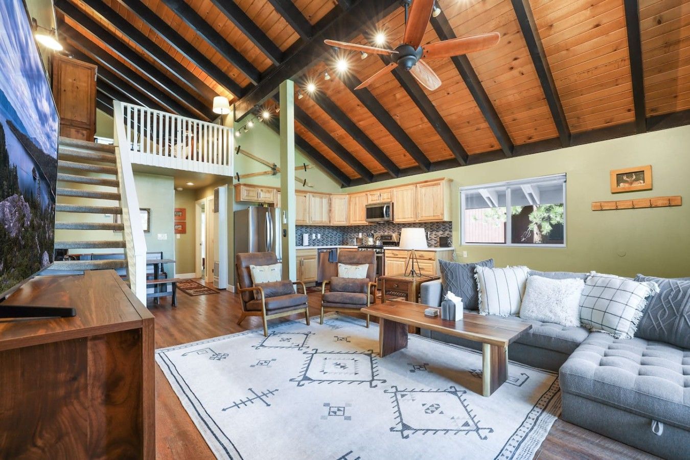 A spacious living area with a high ceiling, wooden beams, a ceiling fan, a dining table, a sofa, and a kitchen in the background.