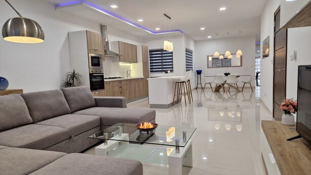 A modern living room with a glass coffee table, a sofa, and a kitchen area with wooden cabinets and a built-in oven. - Carousel - Modern Spacious Apartment  (Qrendi)