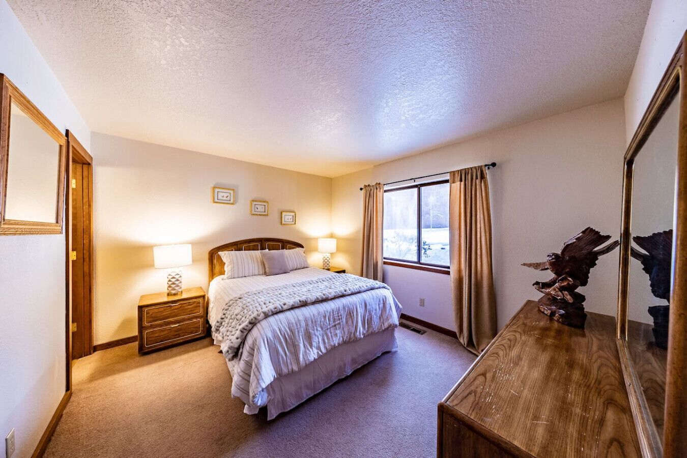 Une chambre à coucher comprenant un lit, des tables de chevet, une commode, un grand miroir et une fenêtre avec des rideaux.