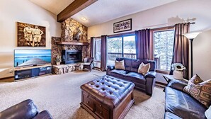A cozy living room with a stone fireplace, leather sofas, a television, and a large window with curtains. - Epic Condo | Beach Access & Meadow Views | Sleeps8 (Zephyr Cove)