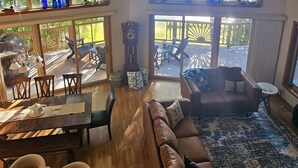 A spacious living room with a large brown leather sofa, a wooden dining table, and a large window overlooking a deck. - Spacious Lakefront Getaway-3BR+Loft, private dock, kayaks/canoe & cozy Fire Pit. (Turtle Lake)