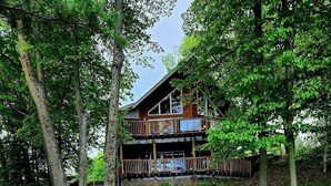 A two-story wooden cabin with a stone staircase leading to the entrance, surrounded by lush greenery. - Spacious Lakefront Getaway-3BR+Loft, private dock, kayaks/canoe & cozy Fire Pit. (Turtle Lake)