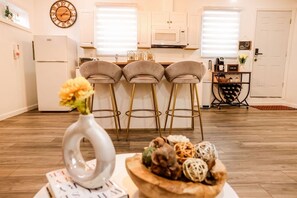 A modern kitchen with a white refrigerator, a microwave, and a coffee machine. There are three bar stools and a wooden table with a bowl of decorative items.
