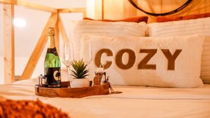 A cozy bedroom with a bed, a pillow with "COZY" written on it, a bottle of champagne, a glass, a small plant, and a wooden tray. - By D&D Vacation Rental (Providence)