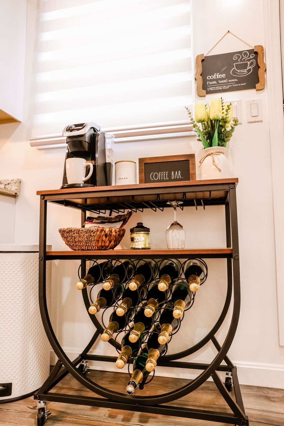 A coffee bar with a sign, a coffee machine, a sugar container, a glass, a basket, and a wine rack with bottles.