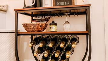 A coffee bar with a sign, a coffee machine, a sugar container, a glass, a basket, and a wine rack with bottles.
