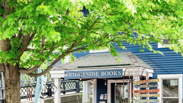 Una fachada con un letrero que dice "Bridgeside Books", rodeada de macetas y un árbol.