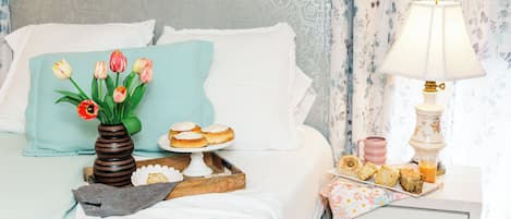 Una cama bien tendida con un jarrón de tulipanes, una bandeja de pasteles y una lámpara en una mesita de noche.