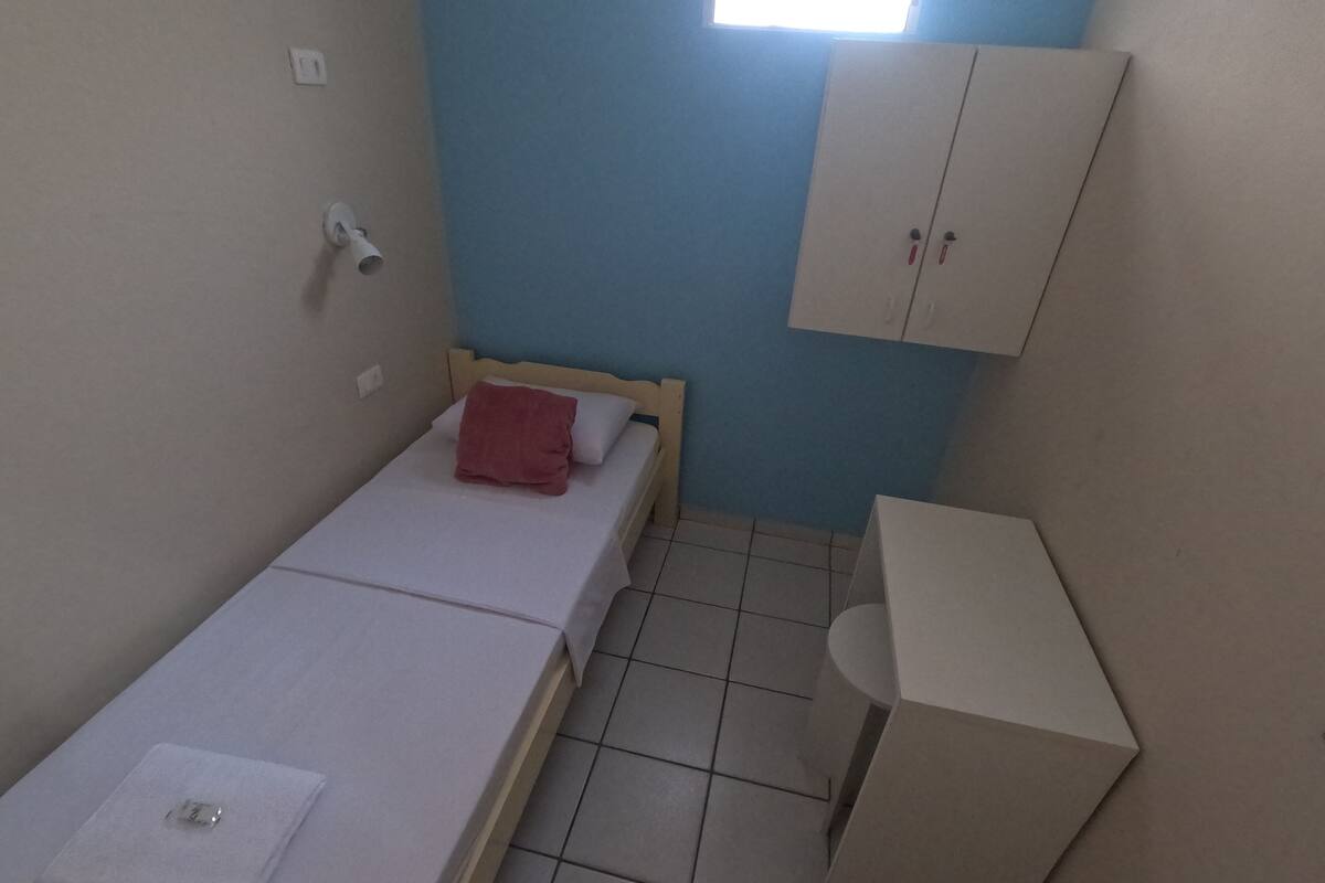 A small, single-bed room with a wooden headboard, a red pillow, and a white blanket. There is a white cabinet with two doors and a small table with a white surface. The room has a tiled floor and a window with a view of the outside.