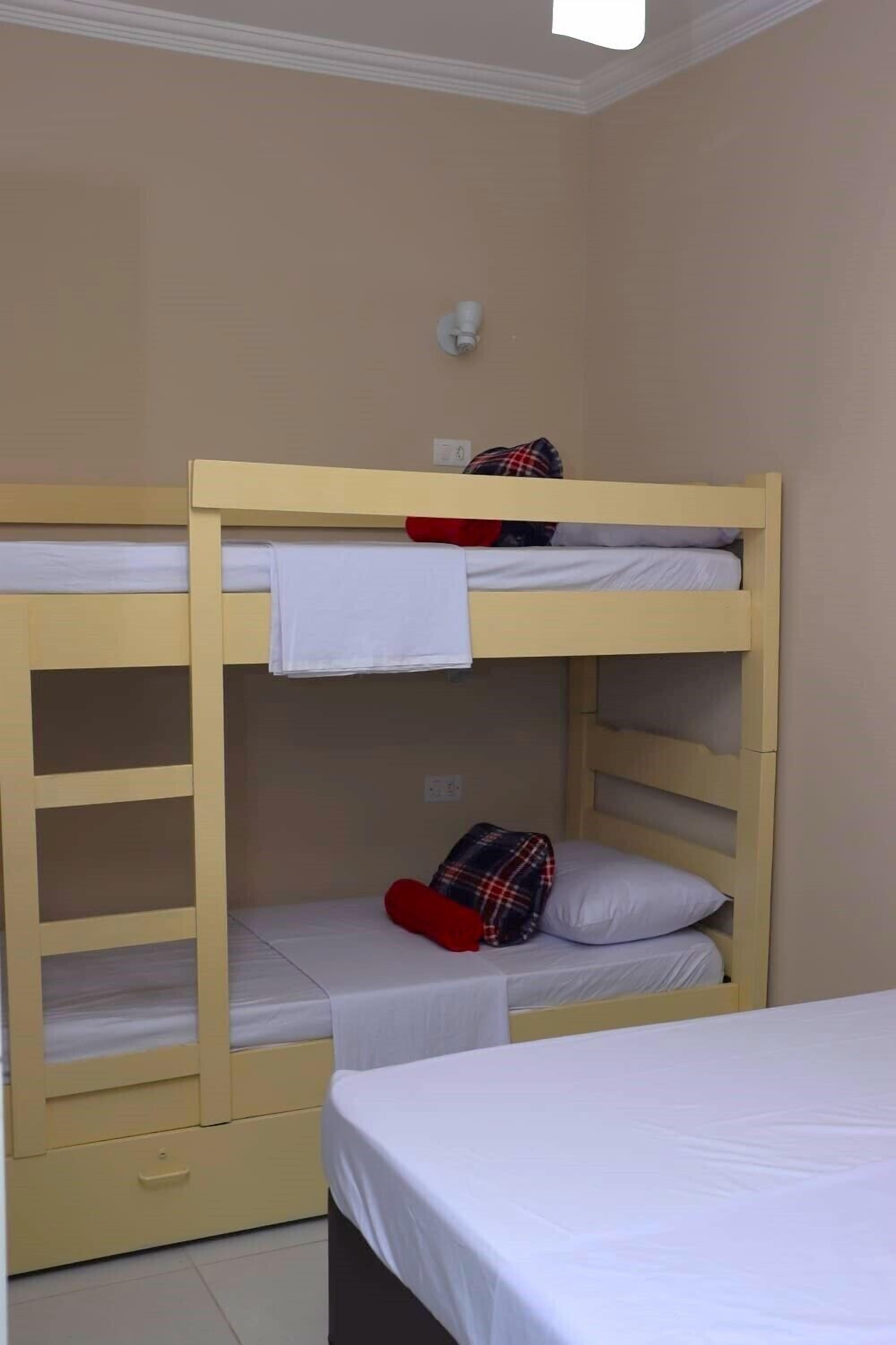 A bunk bed room with a white mattress on the top bunk and a lower bunk with a plaid blanket and a red pillow.