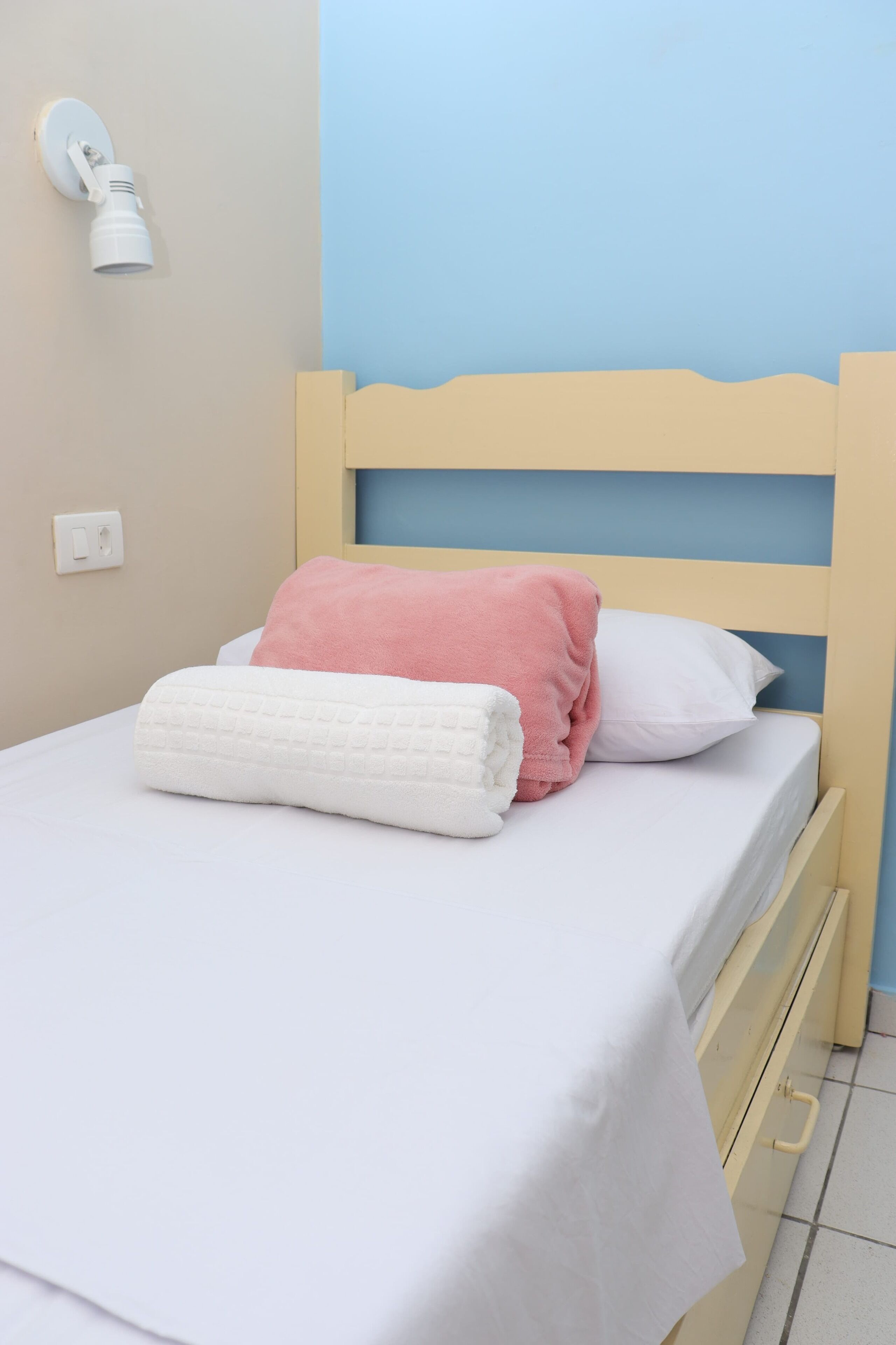 Uma cama com lençol branco, almofada rosa e toalha branca.