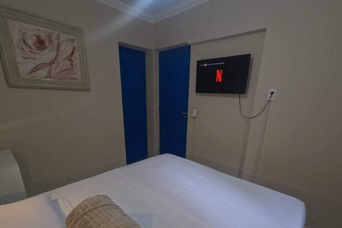 A hotel room with a bed, a rolled towel, a painting, two blue doors, and a TV displaying the Netflix logo.