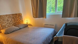 A bedroom with a bed, a nightstand, a heater, a window with curtains, and a chair with a towel. - Maisonnette T2 (Saint-André-les-Vergers)