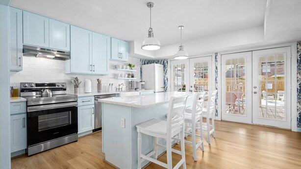 A modern kitchen with light blue cabinets, stainless steel appliances, a central island with bar stools, and a view through glass doors to an outdoor patio.