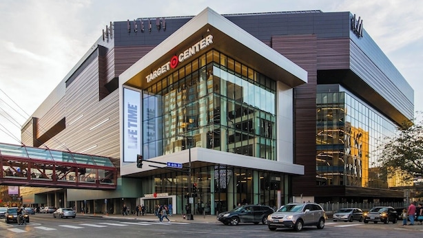 Un bâtiment moderne du Target Center, doté d’une façade en verre, d’un pont piétonnier et offrant une vue sur la rue, avec des voitures et des piétons.