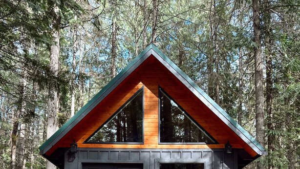 Une cabane dotée d’une terrasse en bois, d’un espace extérieur aménagé et couvert, et d’une grande fenêtre donnant sur la forêt.