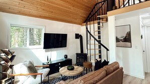 Un vaste espace de vie doté d’un plafond en bois, d’un canapé marron, d’une table basse ronde, d’une bibliothèque et d’un escalier menant à un niveau supérieur.